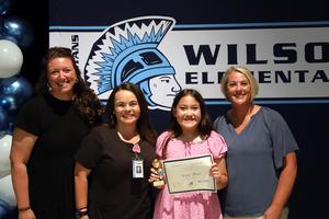 Three women stand with girl in pink dress holding trophy and certificate