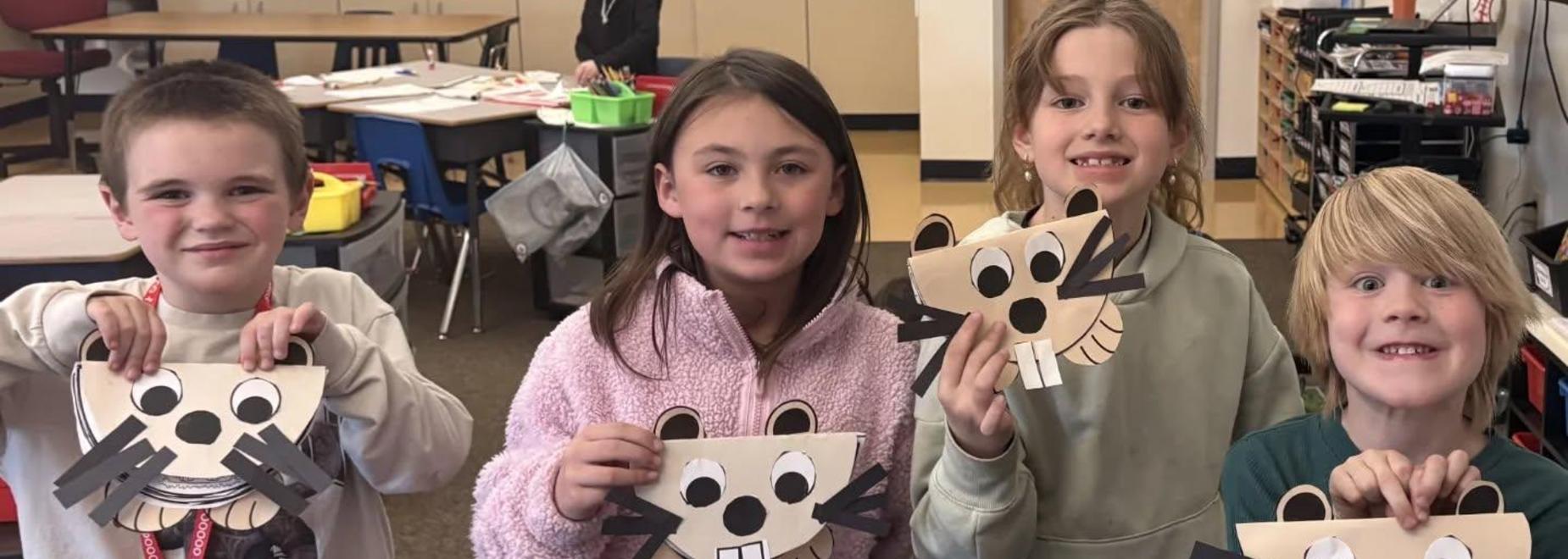 Four children holding handmade beaver masks, smiling at the camera in a classroom setting.