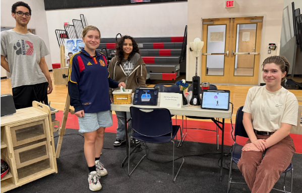 Students stand next to wooden and electronic builds, including the design process on iPads