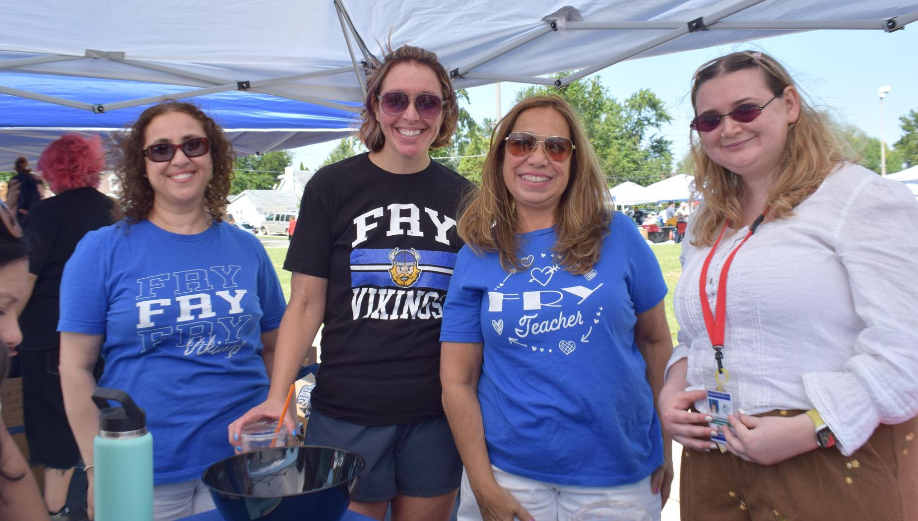 Four women smiling together under a tent, wearing matching blue t-shirts at an event.