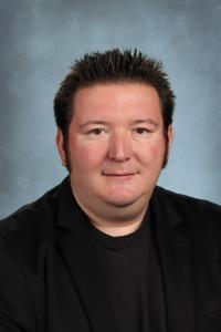Man in a black blazer with short spiky hair and a neutral expression against a blue background.