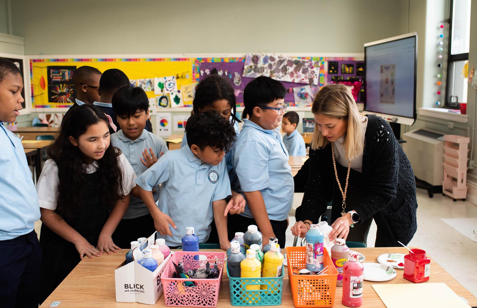Students gather around a table with paints and an instructor in a bright classroom.