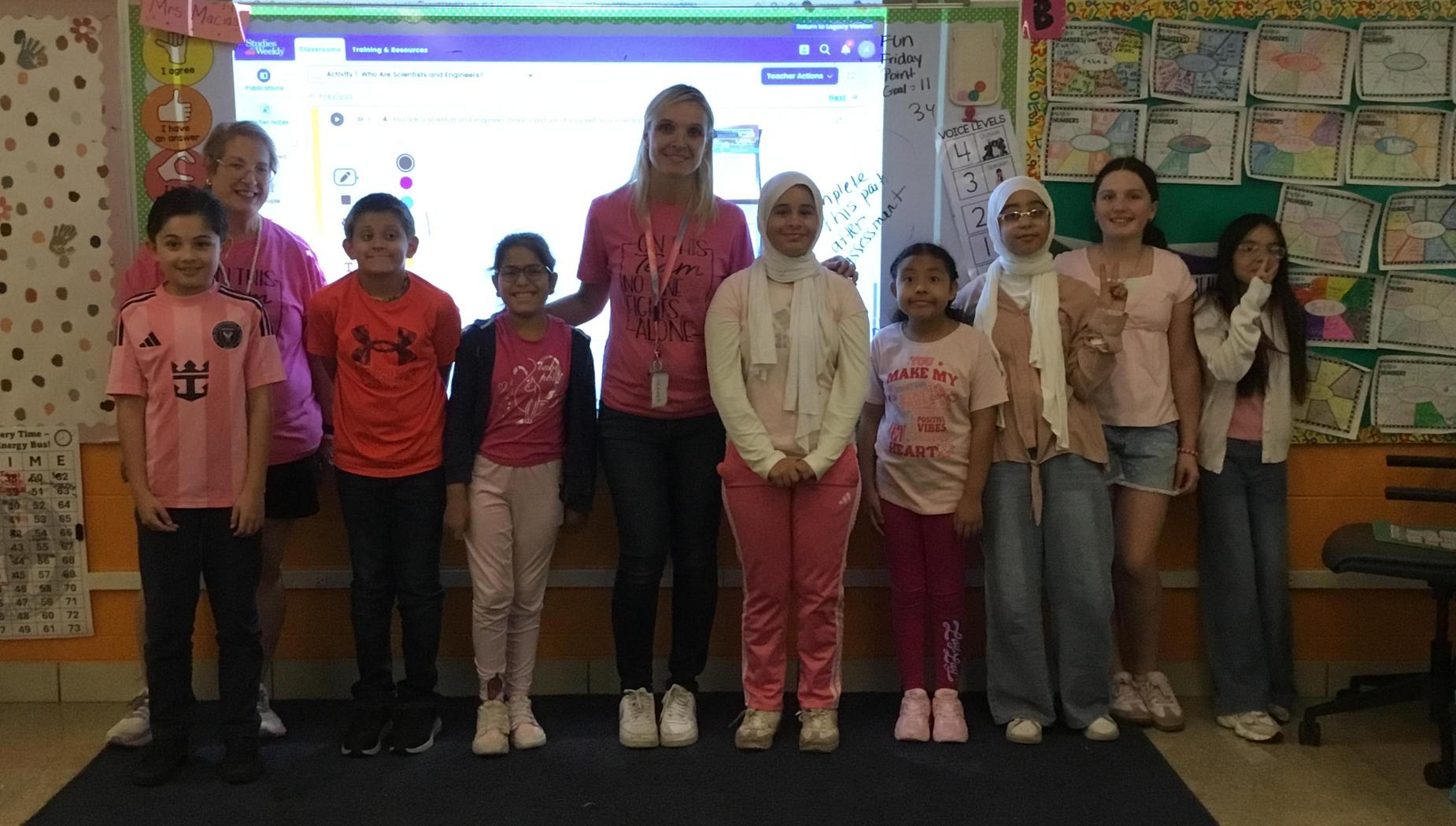 Group of girls and a woman in pink attire standing in a classroom.