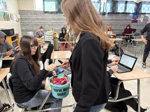 A student shares candy from a basket with another student at a desk.
