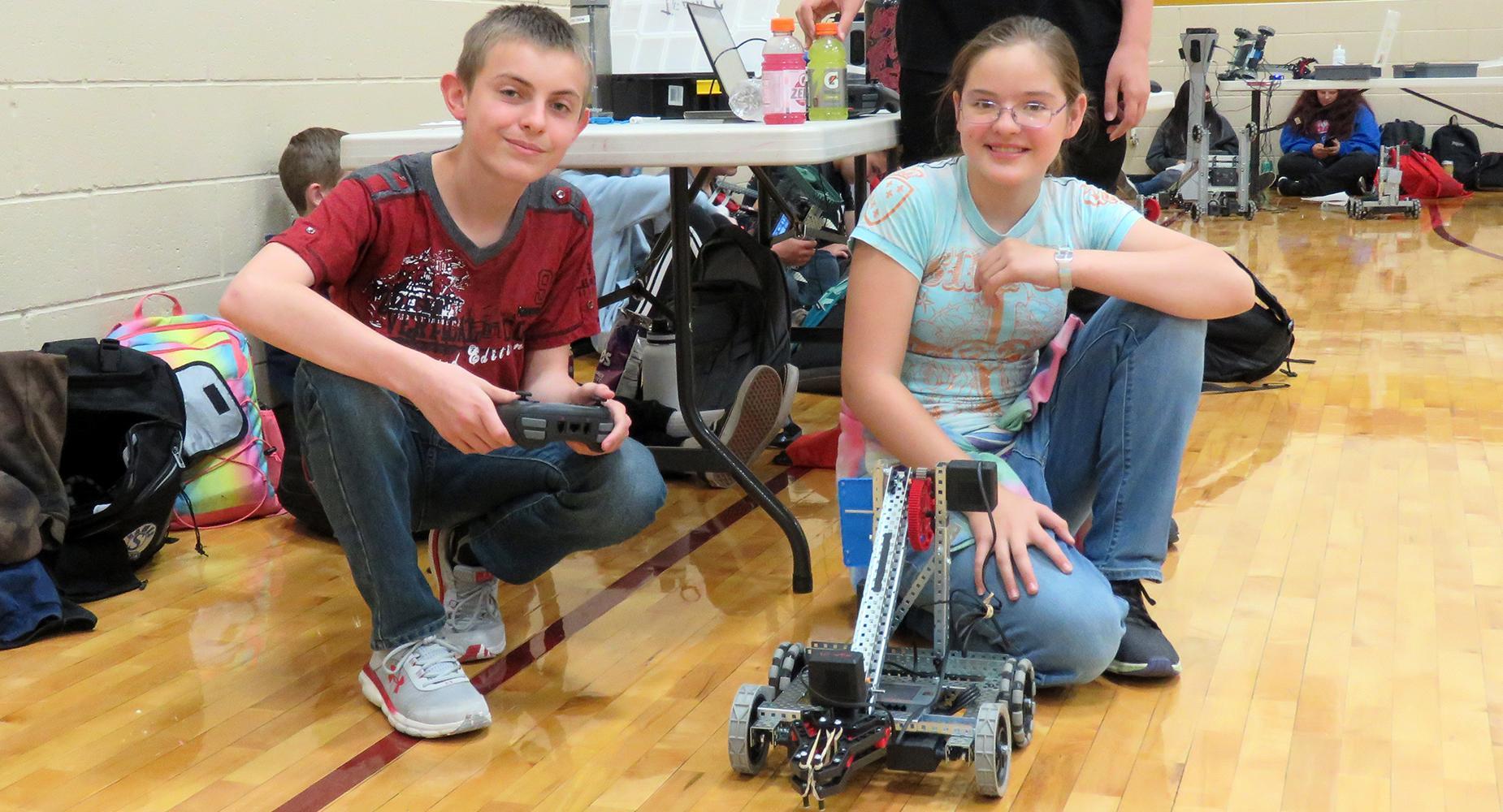 A boy in a red t-shirt and girls in a blue shirt pose with their robot at the East Valley gym.