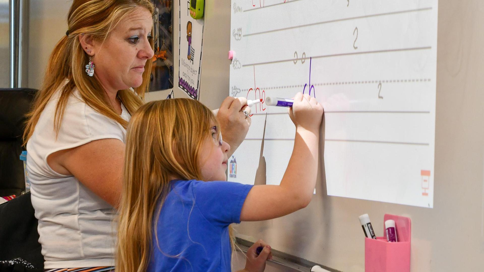 A woman guides a girl in writing on a whiteboard while smiling.