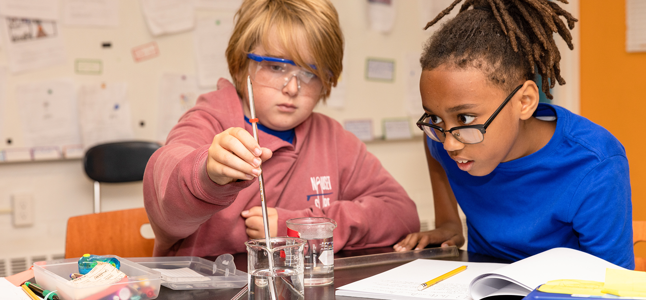 Two students conduct a science experiment in a classroom; one holds a thermometer inside a glass beaker filled with liquid while the other leans in closely to observe