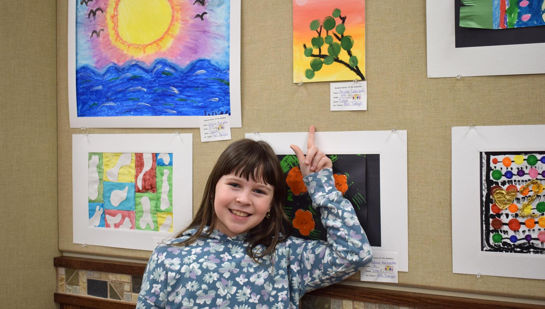 Child smiling next to colorful art displayed on a wall.