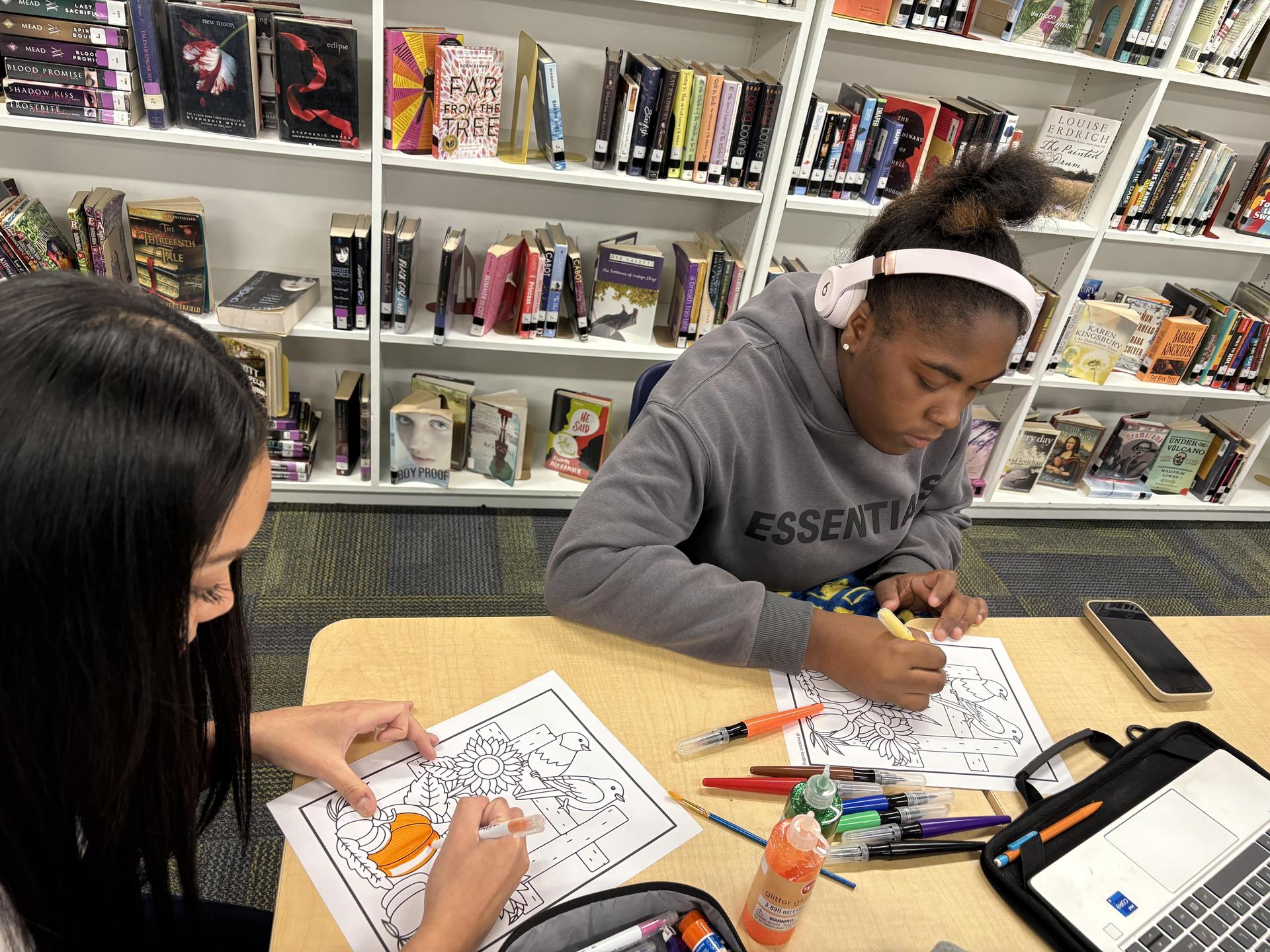 Two students coloring pictures with markers at a table in a library.