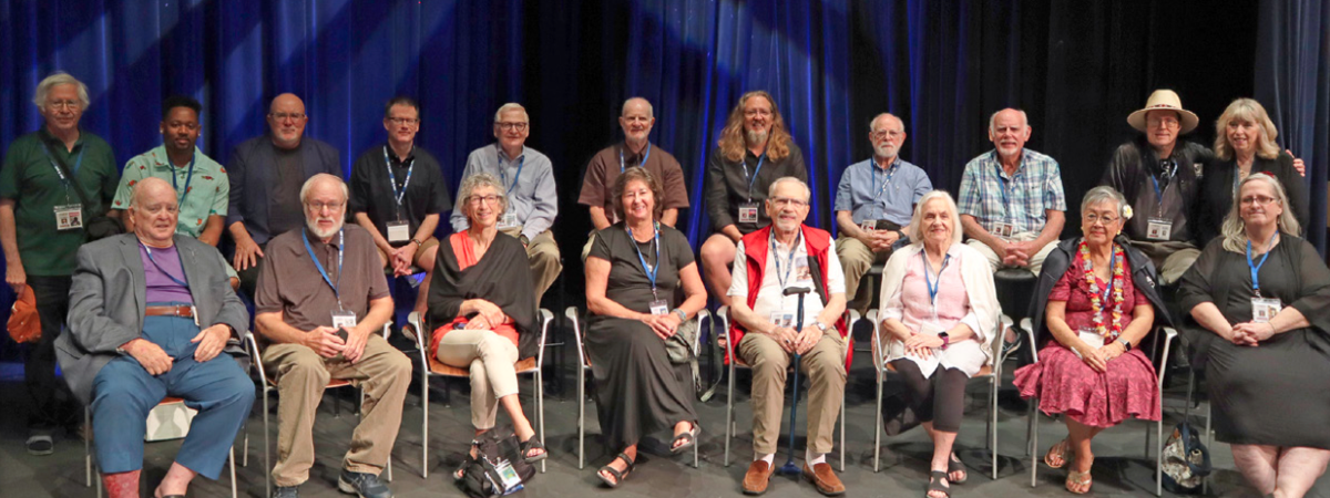 A group photo of participants from the Deaf Authors Book Festival hosted at Texas School for the Deaf (TSD). The group includes authors and attendees seated in two rows on a stage, with a backdrop of deep blue curtains. Many wear name badges and lanyards, smiling warmly for the camera, reflecting a welcoming and inclusive literary event.