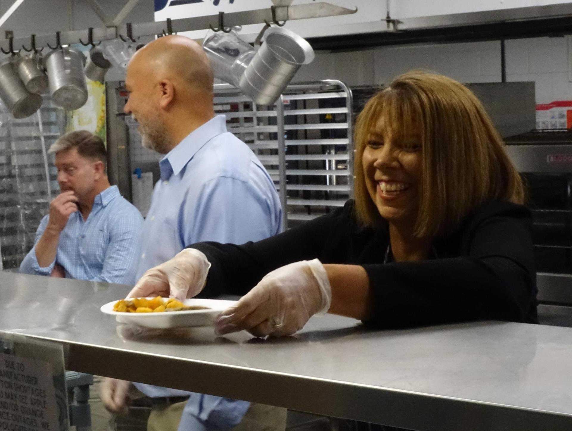 A woman serving food with a smile and two men in the background.