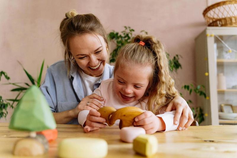 A smiling adult sits with a child playing with wooden toys.