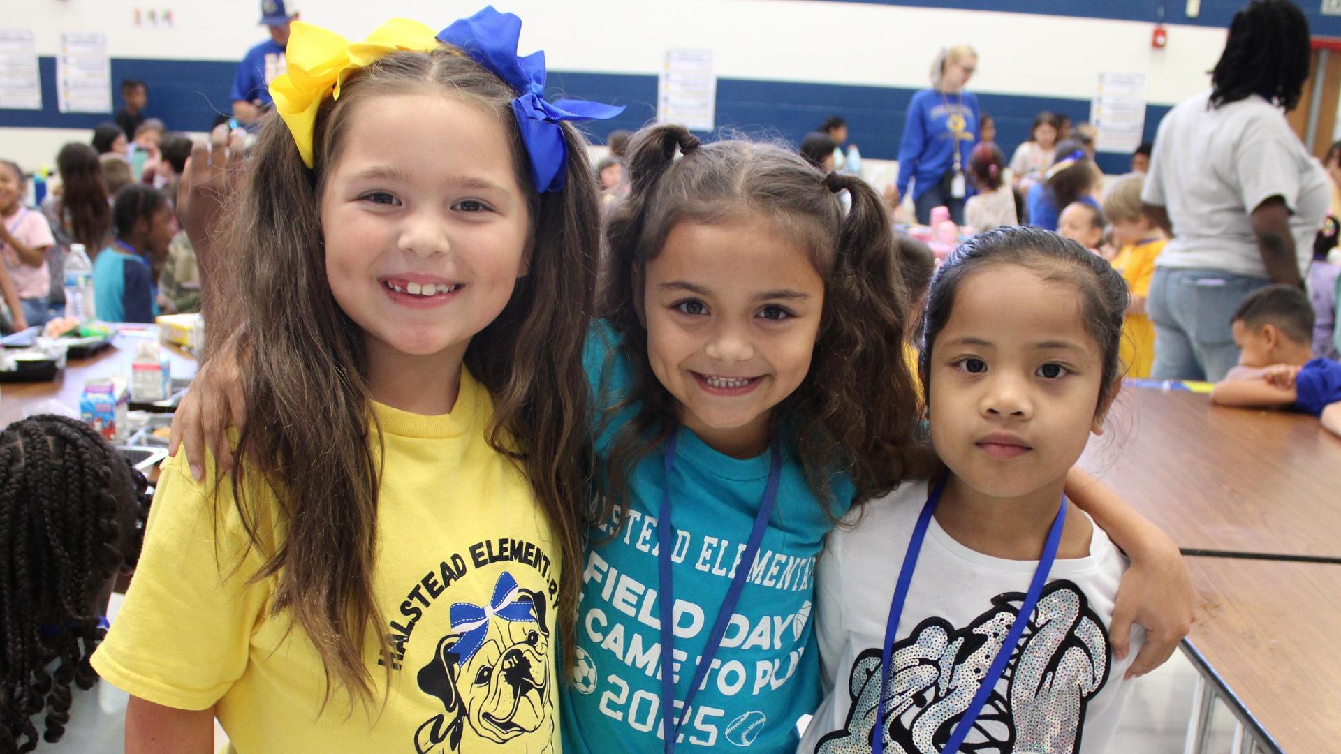 Three girls smiling together, wearing colorful shirts in a school setting.