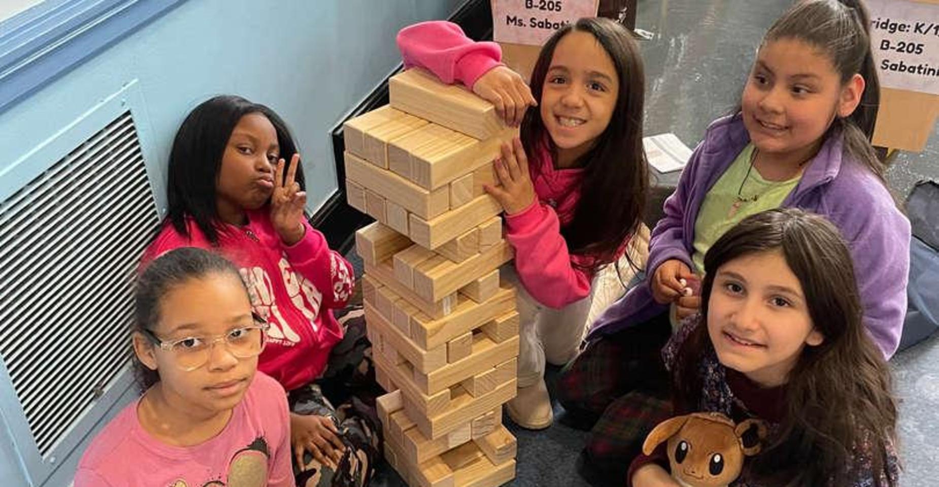 Five children smiling around a tall tower of wooden blocks.