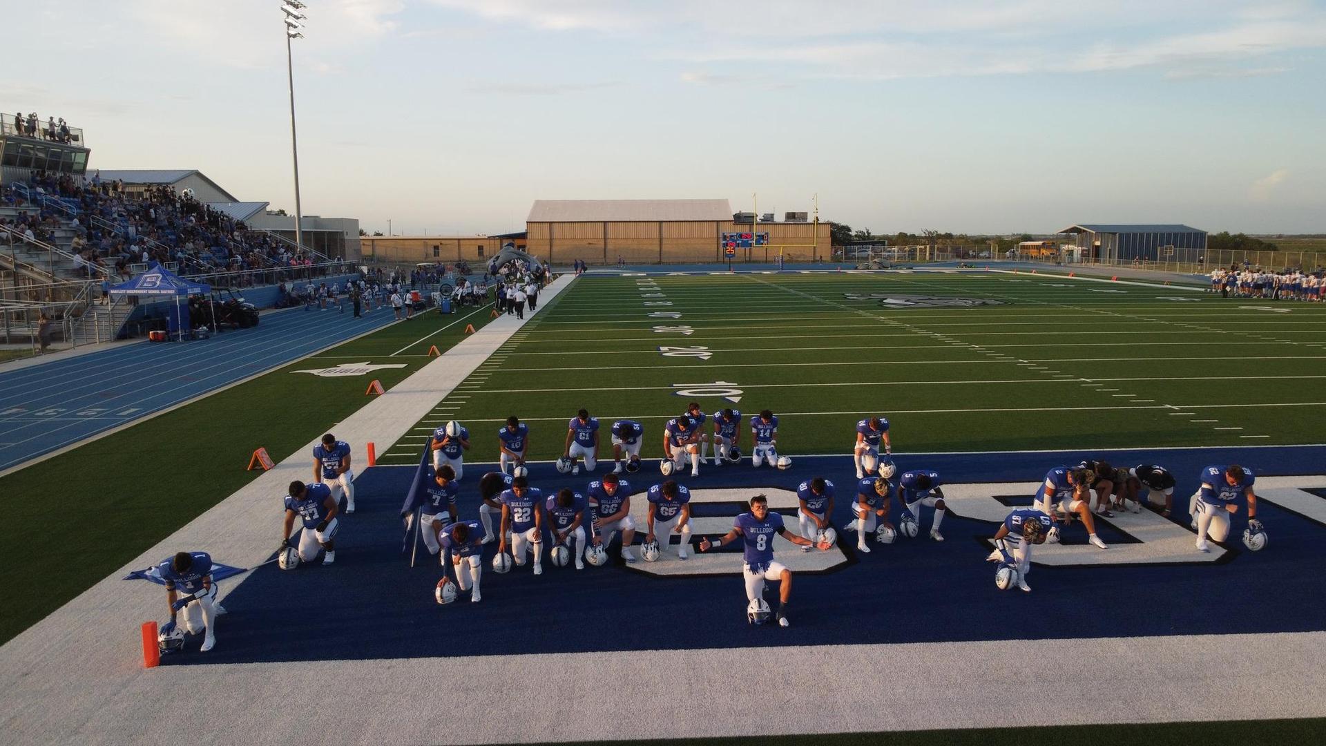 A high school football team kneeling on the field with a bleacher crowd in the background.