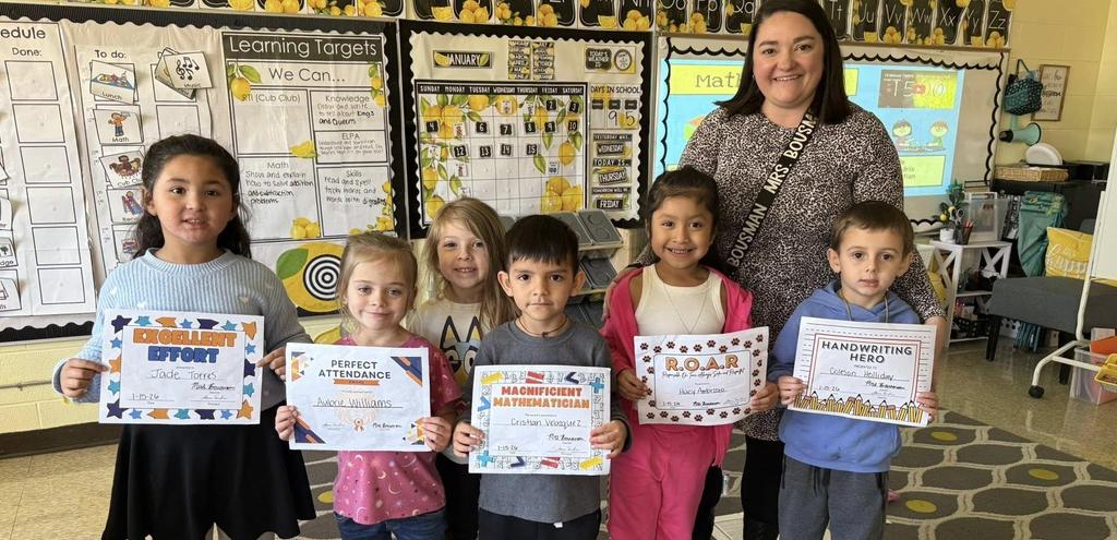 young students standing in a classroom holding certificate awards proudly. adult standing smiling behind them.