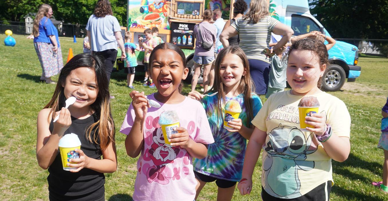 Four children smiling and holding icy treats at an outdoor event.
