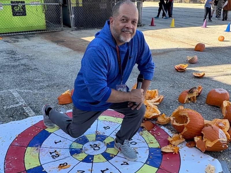 A man kneeling on a dartboard beside scattered pumpkin remnants at an outdoor event.