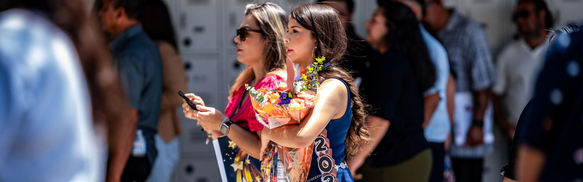 A young woman in a navy dress holds flowers and smiles while walking among a festive crowd.
