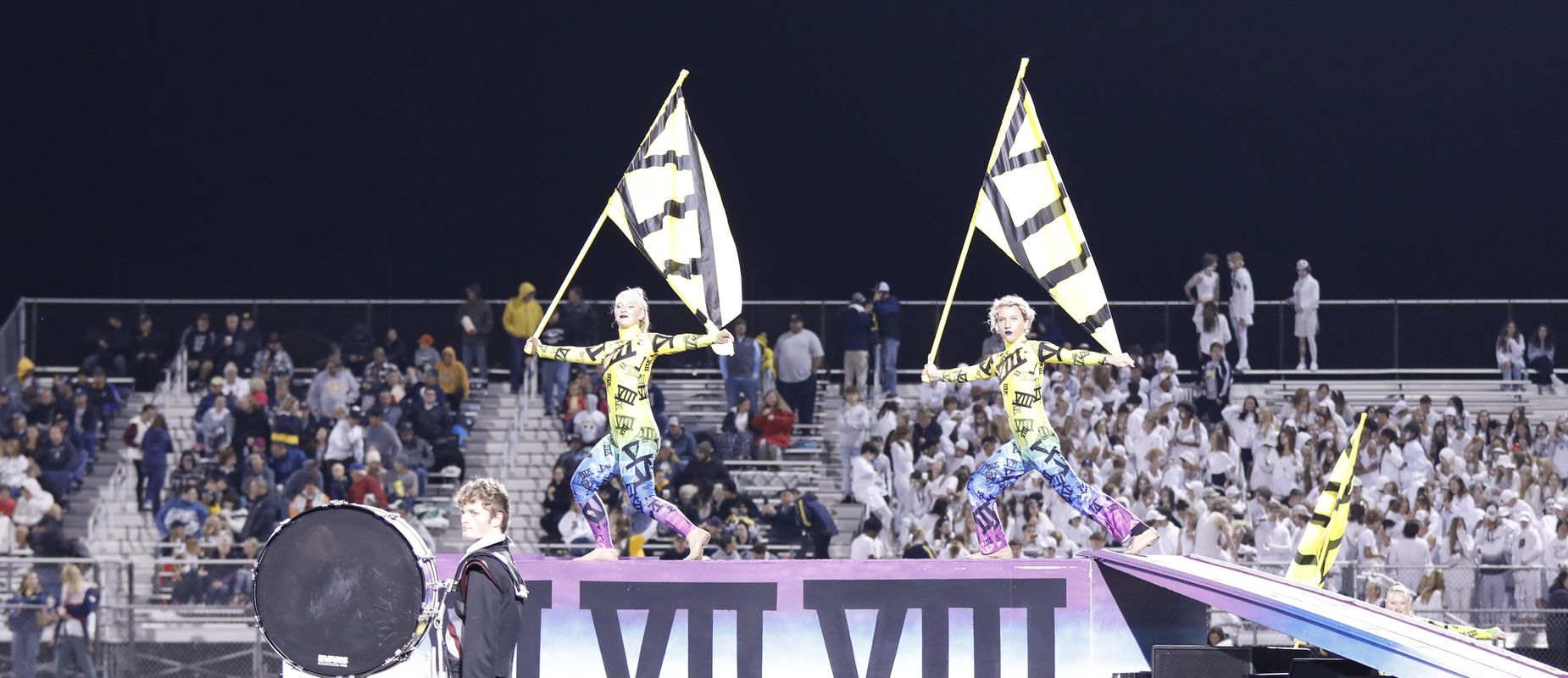 two girls with yellow flags on podium on field with drummer in foreground and audience in stands in background