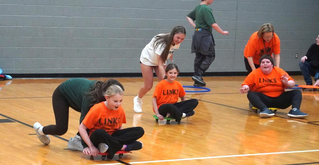Children in orange shirts playing games on scooters in a gymnasium.