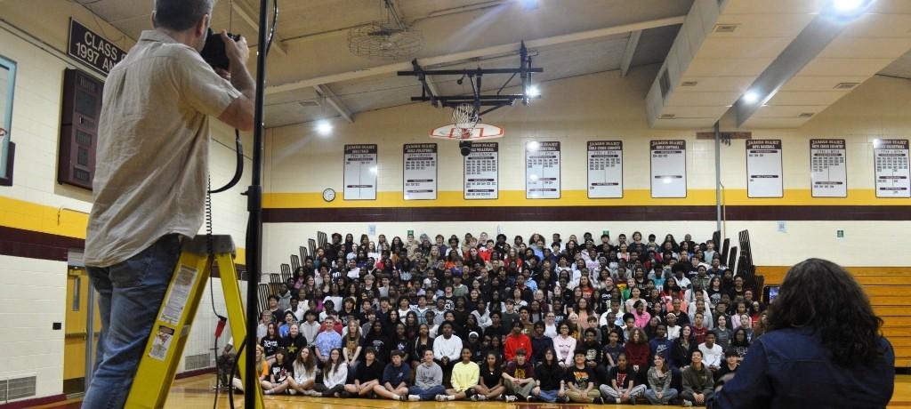 photographer stands on ladder to shoot 8th-grade class seated in gym bleachers