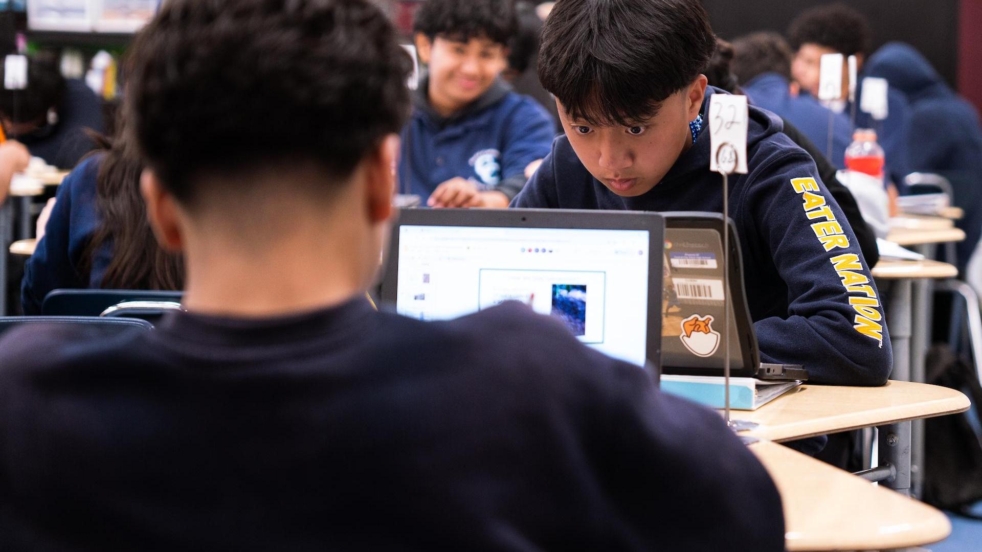 Boys engaged with a laptop in a classroom full of students.