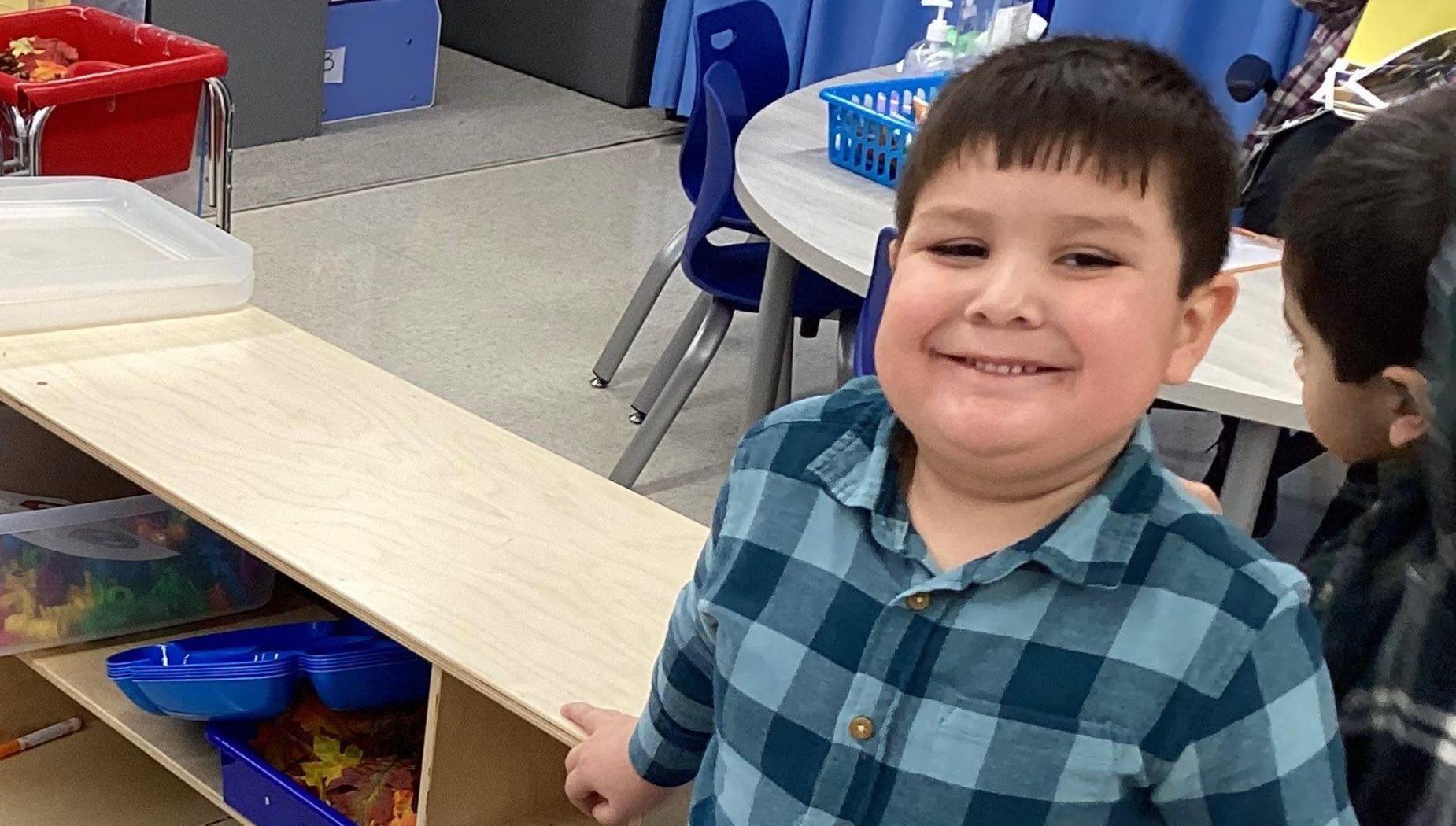 Smiling boy in a plaid shirt next to a wooden shelf in a classroom.