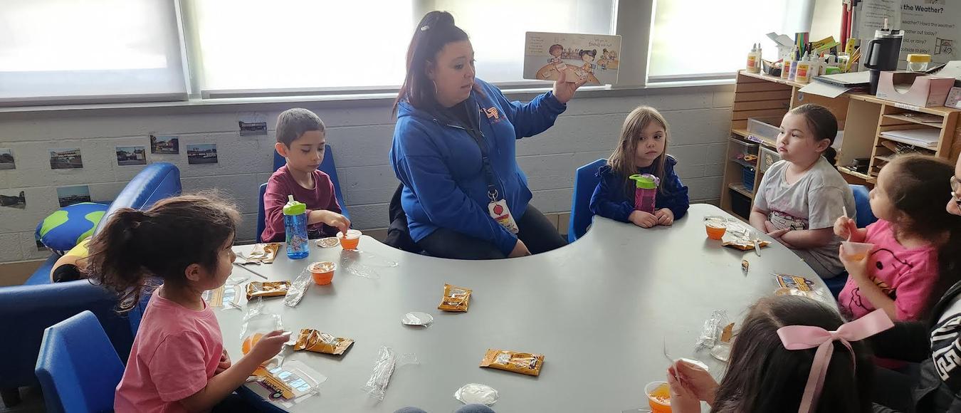 Children are sitting at a table eating fruit and crackers while a teacher is reading them a story.