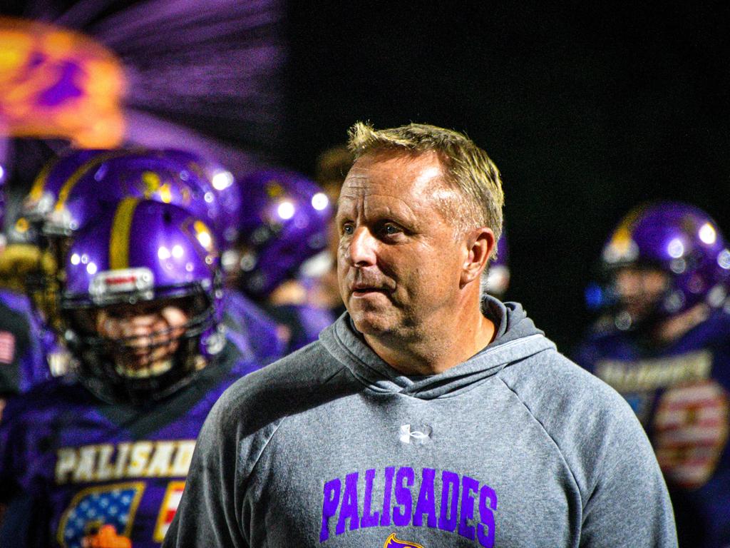 A coach in a grey hoodie stands amidst football players in purple helmets.