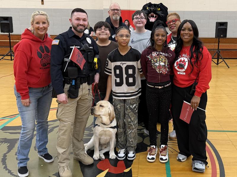 A group of people, including students and an officer, posing with a dog in a gymnasium.