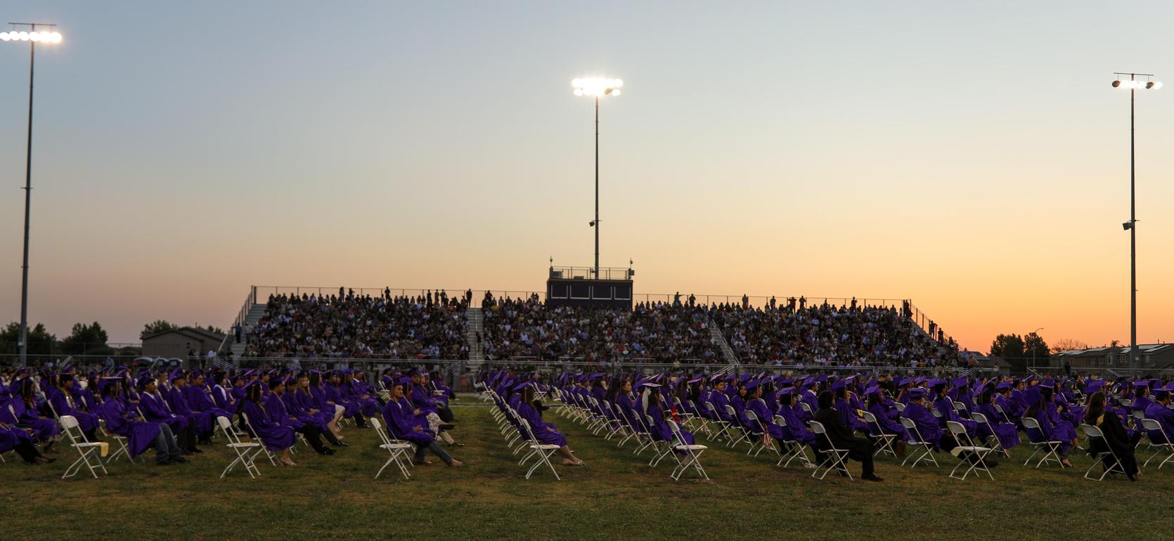 Graduates in purple gowns seated in rows with a large audience in the stands during sunset.