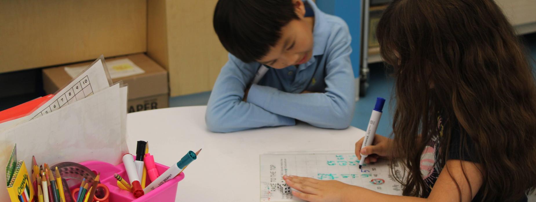A pair of elementary students work together on a white board at a table doing math problems together.