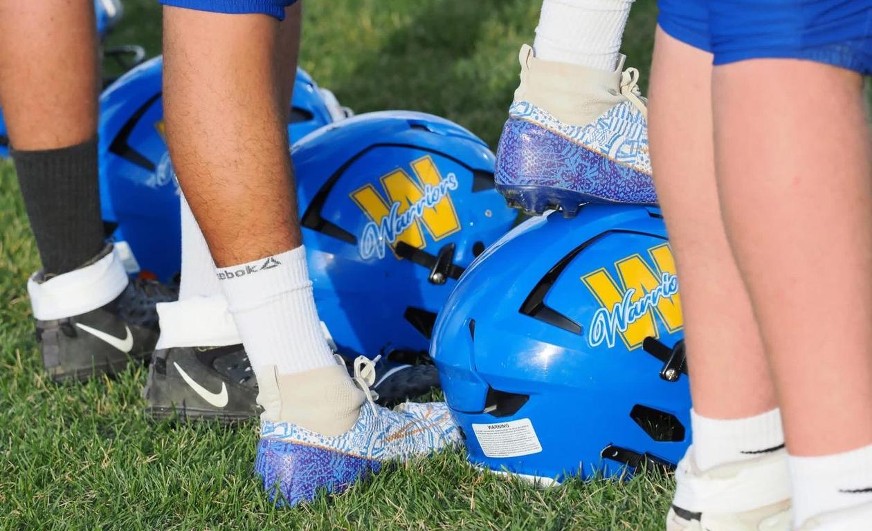 A group of blue football helmets with the 'Warriors' logo and players' feet beside them.