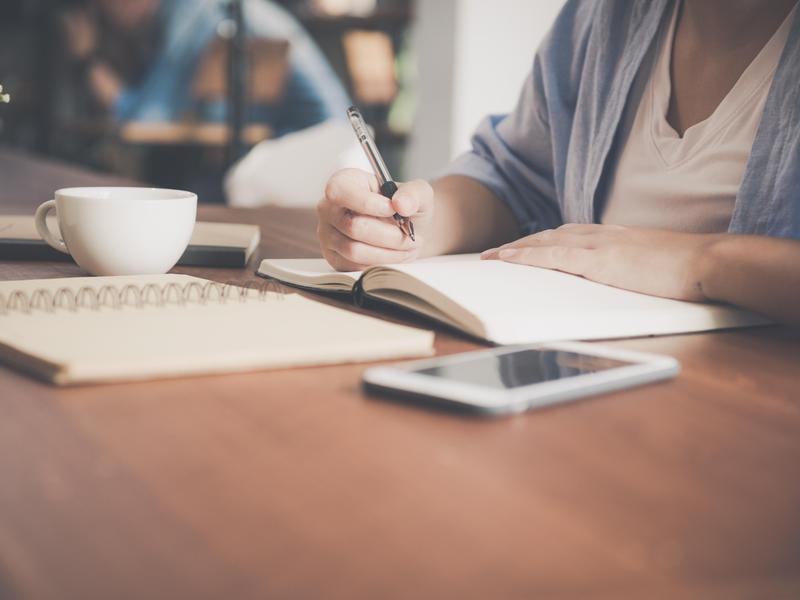 Close-up of a hand writing in a notebook with a cup and phone nearby.