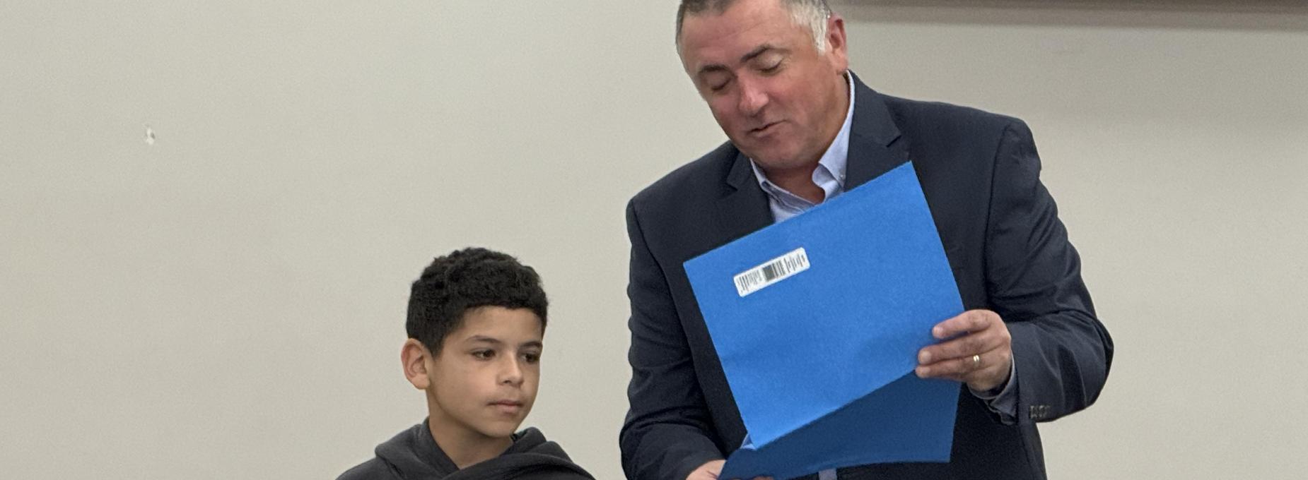 A man shows a folder to a boy as they stand together before an audience.