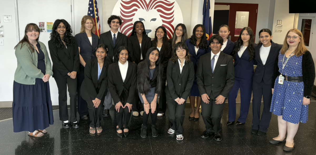 Students stand in business dress with advisors in the high school lobby, next to an American flag