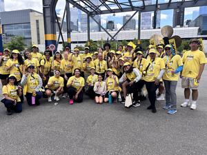 A large group of students and staff wearing matching yellow Race of Hope shirts pose together outdoors after completing a race. Many participants are smiling and holding up medals, with a cityscape and event structures visible in the background, conveying a sense of community, accomplishment, and support for mental health awareness.