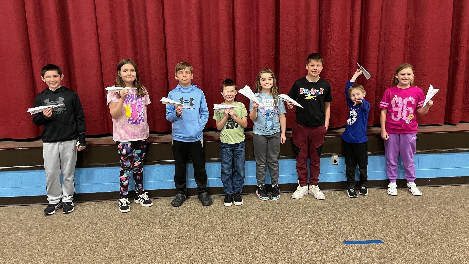Eight children holding paper airplanes, standing in a line on a stage.