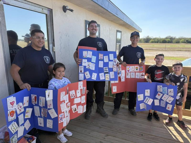 3 Firefighters dressed in blue standing with 2 boys and a girl holding large red and blue thank you cards