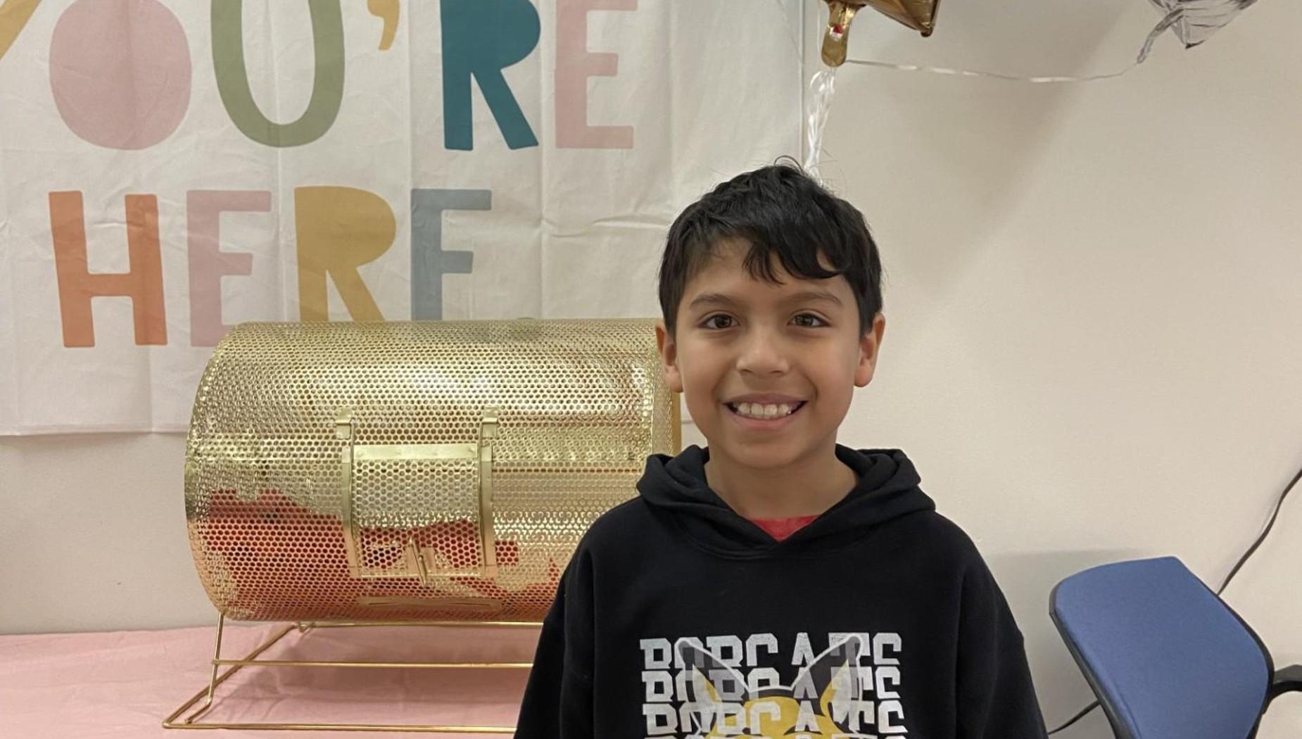 A smiling child stands in front of a gold raffle drum and colorful banner.