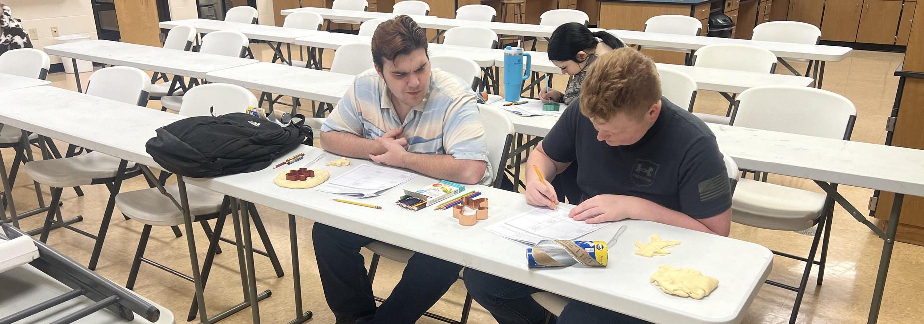 Two students working at a table with papers and snacks in a classroom setting.