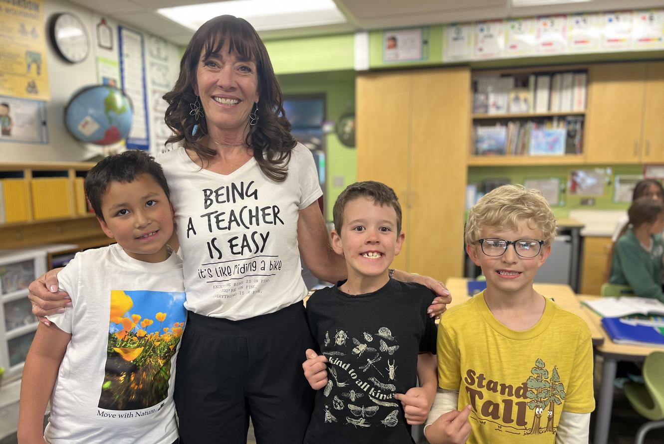 A teacher poses with four smiling children, all wearing colorful t-shirts.