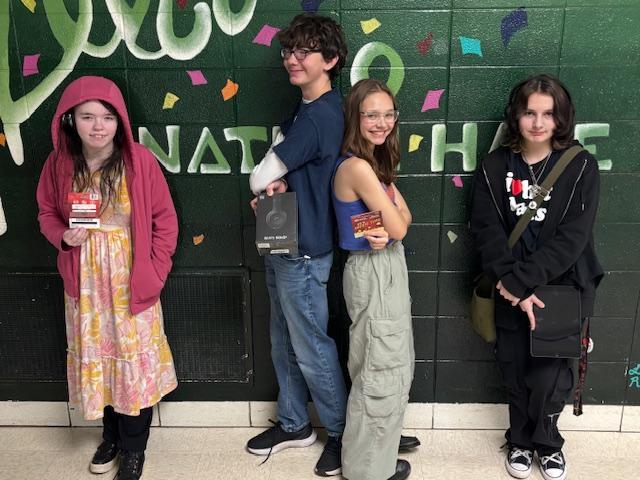 Four teenagers pose in front of a colorful mural, holding cards and smiling.