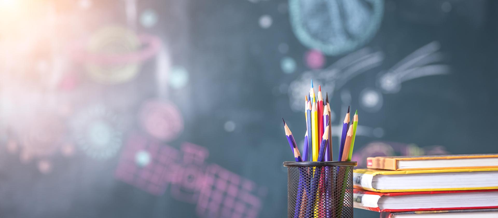 Colorful pencils in a holder with stacked books and a chalkboard background.
