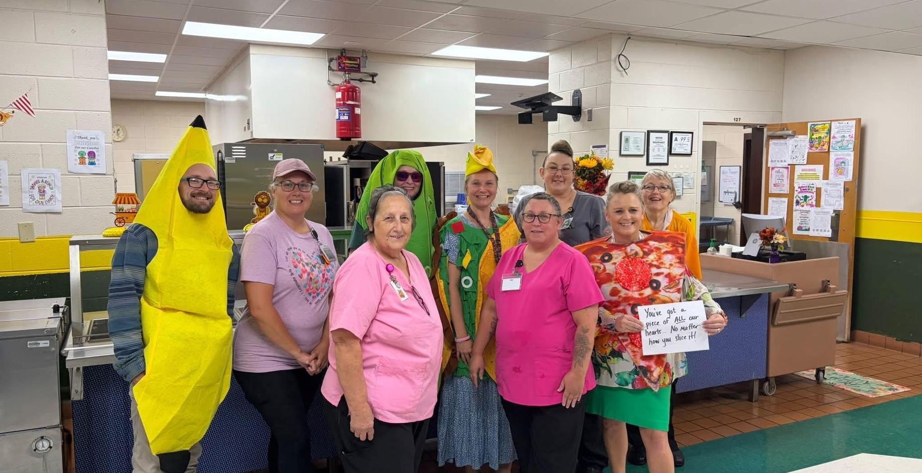 Group of staff in costumes posing in a cafeteria setting.