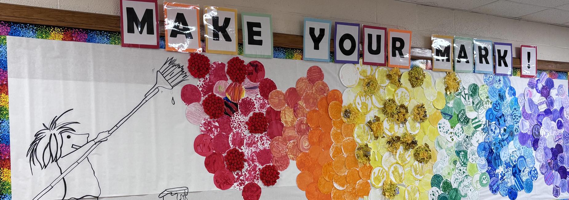 The entryway bulletin board at MES shows various colored and designed paper plates made by students. The caption reads "make your mark!"