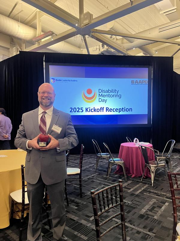 Image depicts Mr. Brindza in a gray suit while holding his award at the event.