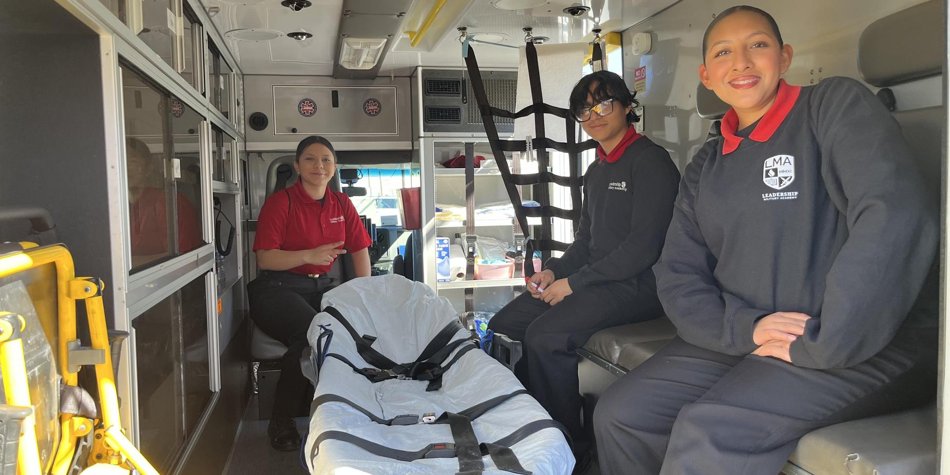 Three individuals in an ambulance, smiling and seated near a stretcher.