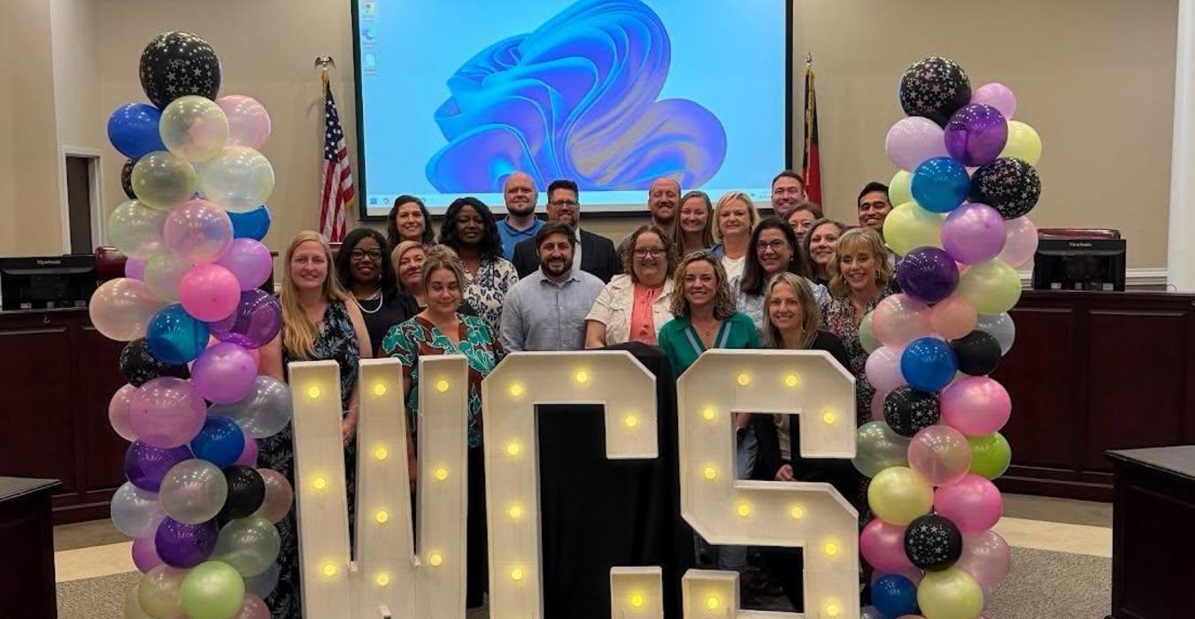 Group of people posing with large illuminated letters 'WCS' and colorful balloons.
